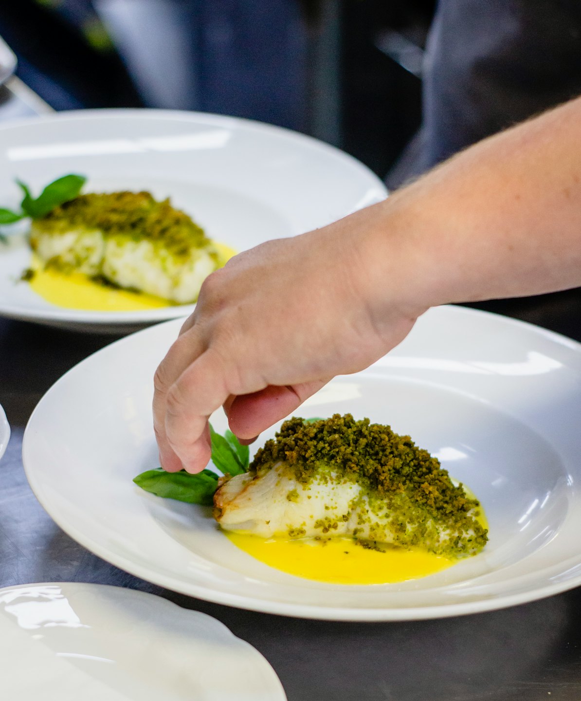 Chef plating a white fish dish with basil on yellow sauce.